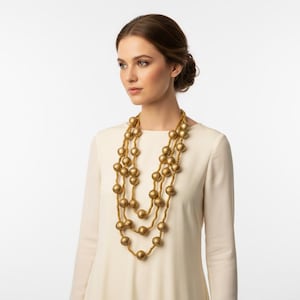 May include: A three-strand gold-tone necklace with large, round beads. The necklace is worn over a cream-colored long-sleeved dress. The background is plain white, highlighting the jewelry.