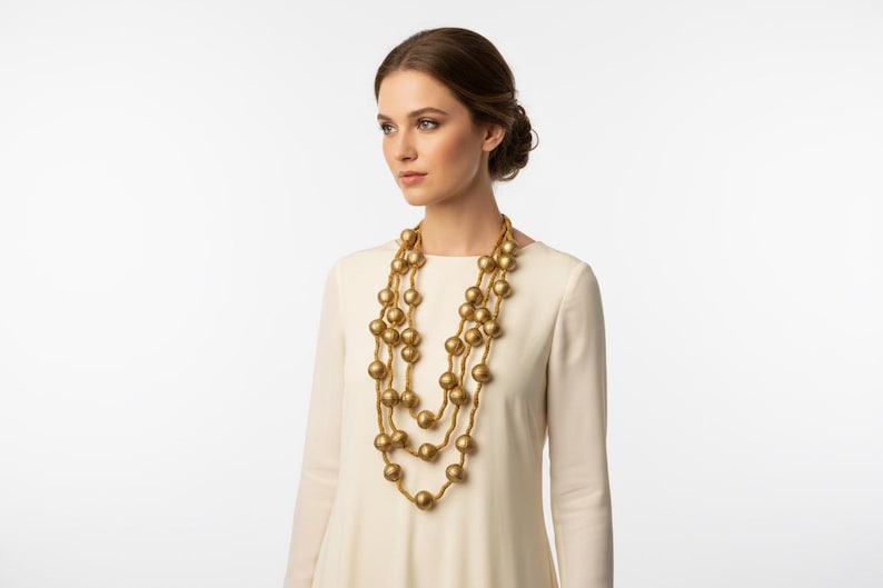 May include: A three-strand gold-tone necklace with large, round beads. The necklace is worn over a cream-colored long-sleeved dress. The background is plain white, highlighting the jewelry.