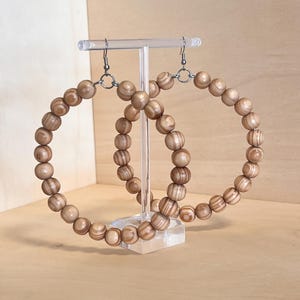 May include: A pair of large hoop earrings made of light brown wooden beads. The beads are strung together to form a circular shape, with silver-colored hooks. The earrings are displayed on a clear acrylic stand.