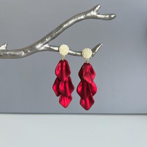 May include: A pair of red petal-shaped earrings with gold-colored circular studs. The earrings are displayed on a silver-colored branch against a gray background. The earrings have a layered design, with each earring featuring three cascading petals.