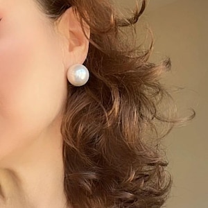 May include: A large, round, white pearl stud earring is visible on the ear. The earring is a prominent feature against the backdrop of brown hair and a navy blue top. The image is a close-up shot.