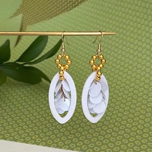 Puede incluir: Pendientes ovalados blancos con cuentas doradas y lentejuelas blancas. Los pendientes tienen un diseño colgante.