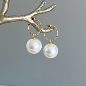 May include: A pair of gold hoop earrings featuring large, round, white faux pearls suspended from the bottom of each thin gold wire hoop. The earrings include gold post-style backings and exhibit a smooth, reflective surface on both the metal and the pearl components.