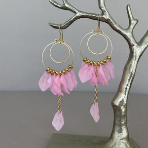 May include: Pink flower petal earrings with gold hoops and a dangling chain. The earrings are hanging from a silver tree branch.