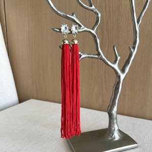 Può includere: Un paio di orecchini a nappa rossi con dettagli in strass trasparenti. Gli orecchini sono esposti su un porta orecchini a forma di albero argentato.