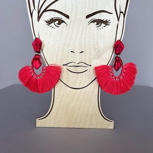 May include: Red tassel earrings with a gold ring and a red satin bow. The earrings are on a wooden mannequin head.