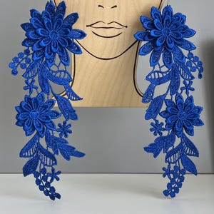 May include: A pair of royal blue lace earrings. Each earring features a large flower at the top, with cascading floral and leaf details. The earrings are displayed against a wooden cutout of a face.