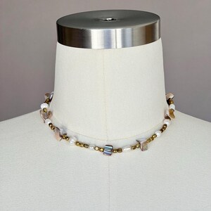 May include: A delicate necklace featuring a mix of white pearls, gold beads, and iridescent shell pieces. The necklace is displayed on a white mannequin.
