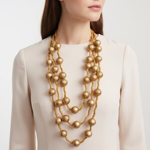 May include: A three-strand gold-tone necklace with large, round beads. The necklace is layered and hangs against a cream-colored dress. The beads have a matte finish and are strung on a thin, tan cord. The necklace is a statement piece.