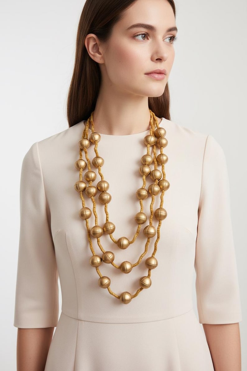 May include: A three-strand gold-tone necklace with large, round beads. The necklace is layered and hangs against a cream-colored dress. The beads have a matte finish and are strung on a thin, tan cord. The necklace is a statement piece.