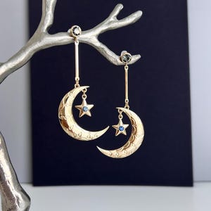 Può includere: Orecchini dorati con lune crescenti e stelle pendenti con accenti blu. Gli orecchini sono sospesi a un ramo color argento su uno sfondo scuro. Le lune hanno una superficie strutturata.