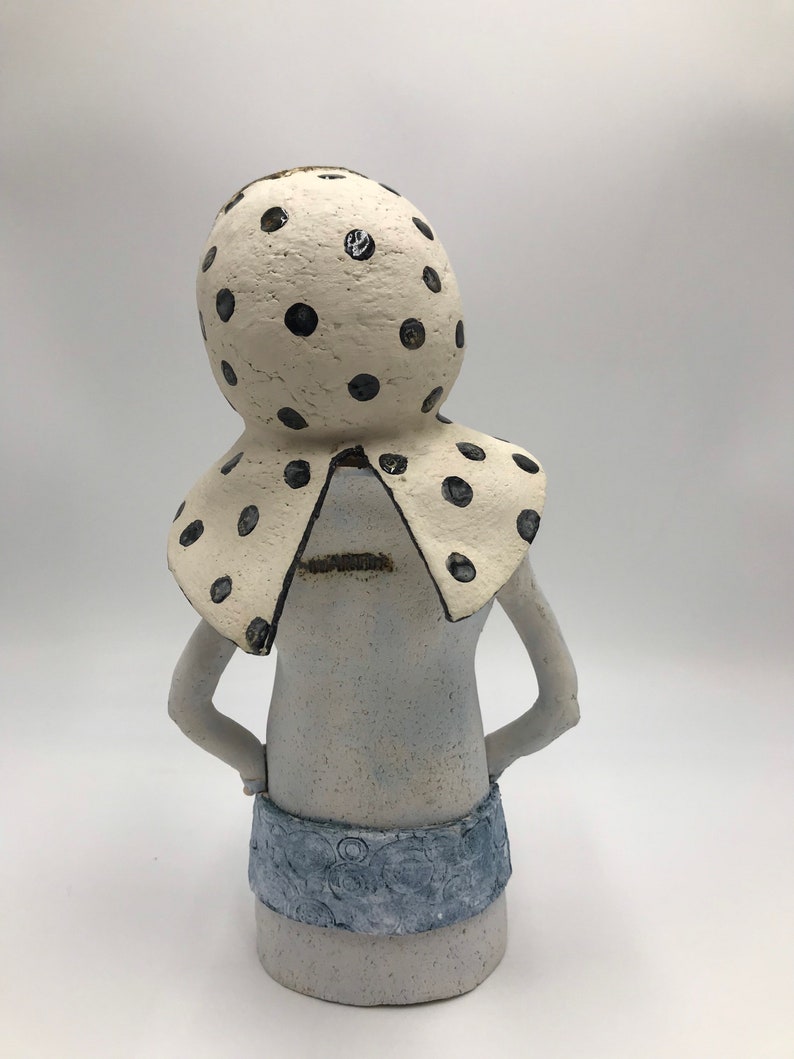 K&ouml;nnte beinhalten: Eine wei&szlig;e Keramikfigur einer Person mit einem gro&szlig;en, wei&szlig;en, gepunkteten Hut und einem blauen, gemusterten Rock.