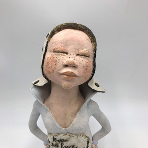 K&ouml;nnte beinhalten: Eine Keramikfigur einer Person mit geschlossenen Augen, die einen wei&szlig;en Hut mit schwarzen Punkten und ein wei&szlig;es Hemd mit einem blau-wei&szlig; gemusterten Rock tr&auml;gt. Die Figur hat ein wei&szlig;es Schild um die Taille mit handschriftlichem Text.