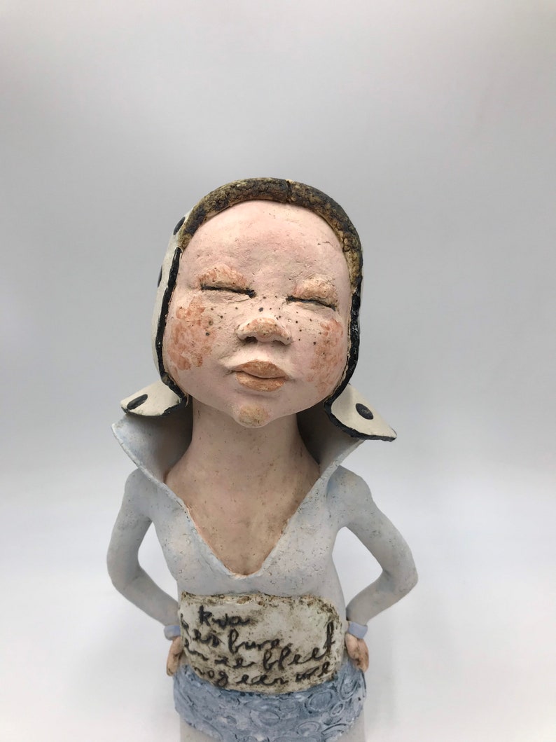 K&ouml;nnte beinhalten: Eine Keramikfigur einer Person mit geschlossenen Augen, die einen wei&szlig;en Hut mit schwarzen Punkten und ein wei&szlig;es Hemd mit einem blau-wei&szlig; gemusterten Rock tr&auml;gt. Die Figur hat ein wei&szlig;es Schild um die Taille mit handschriftlichem Text.