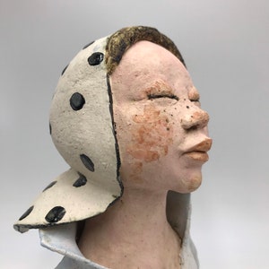 K&ouml;nnte beinhalten: Eine Keramikskulptur eines Menschenkopfes und der Schultern. Die Person tr&auml;gt einen wei&szlig;en Hut mit schwarzen Punkten und ein hellblaues Hemd. Die Augen der Person sind geschlossen und der Mund leicht ge&ouml;ffnet.