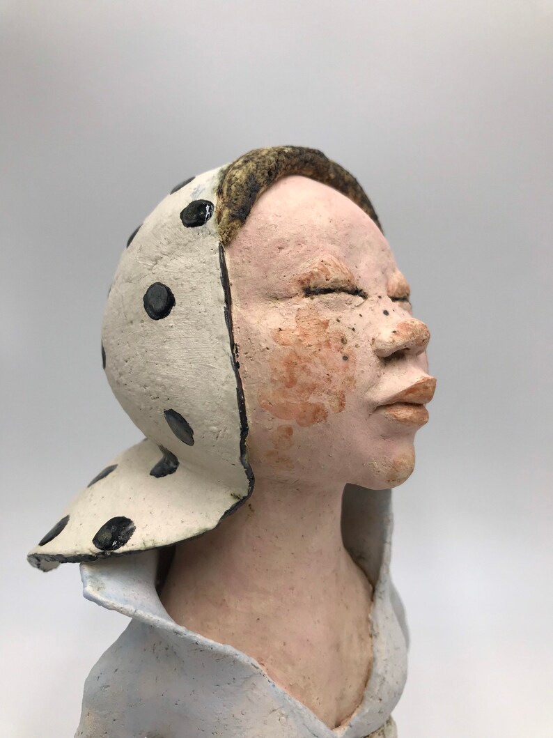 K&ouml;nnte beinhalten: Eine Keramikskulptur eines Menschenkopfes und der Schultern. Die Person tr&auml;gt einen wei&szlig;en Hut mit schwarzen Punkten und ein hellblaues Hemd. Die Augen der Person sind geschlossen und der Mund leicht ge&ouml;ffnet.