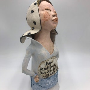 K&ouml;nnte beinhalten: Eine Keramikfigur einer Frau mit einem wei&szlig;en Kopftuch mit schwarzen Punkten, die ein hellblaues Hemd und einen Rock tr&auml;gt. Die Figur hat eine handschriftliche Inschrift auf ihrer Brust, die lautet "Kva er so furtig en ze ble nog en en".