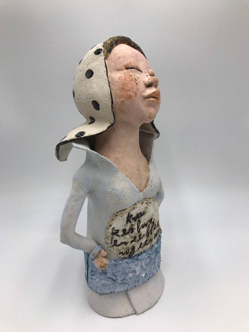 K&ouml;nnte beinhalten: Eine Keramikfigur einer Frau mit einem wei&szlig;en Kopftuch mit schwarzen Punkten, die ein hellblaues Hemd und einen Rock tr&auml;gt. Die Figur hat eine handschriftliche Inschrift auf ihrer Brust, die lautet "Kva er so furtig en ze ble nog en en".