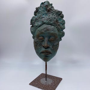 Op de afbeelding: Een bronzen sculptuur van een gezicht met gesloten ogen en getextureerd haar. De sculptuur heeft een groen-koperen patina en is gemonteerd op een metalen standaard met een vierkante basis. Het gezicht heeft geëtste details.