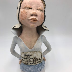K&ouml;nnte beinhalten: Eine Keramikfigur einer Person mit geschlossenen Augen, die einen wei&szlig;-schwarz gemusterten Hut und ein hellblaues Hemd tr&auml;gt. Die Figur hat ein wei&szlig;es Schild um die Taille mit dem Text "Kwas een brug bleef ze bleef nog een uur".