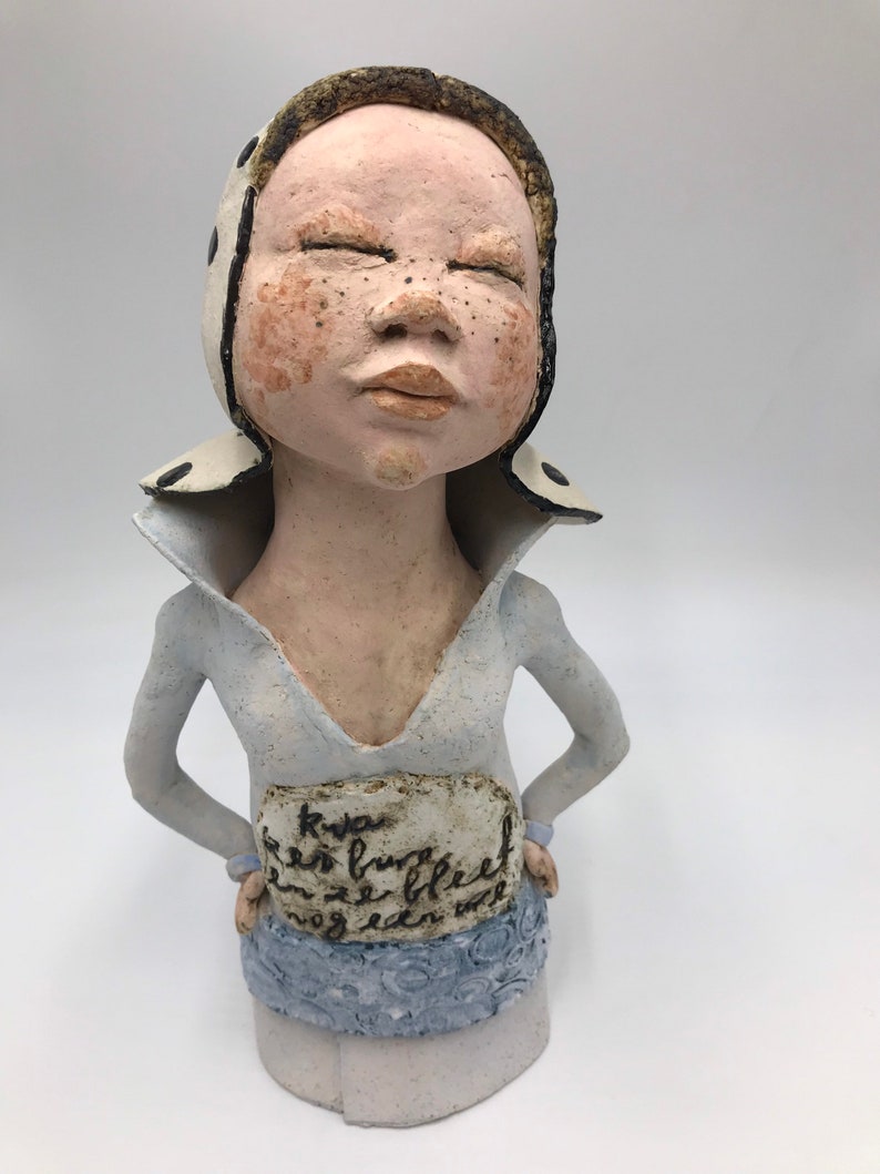K&ouml;nnte beinhalten: Eine Keramikfigur einer Person mit geschlossenen Augen, die einen wei&szlig;-schwarz gemusterten Hut und ein hellblaues Hemd tr&auml;gt. Die Figur hat ein wei&szlig;es Schild um die Taille mit dem Text "Kwas een brug bleef ze bleef nog een uur".