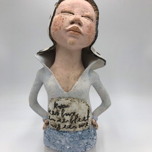 K&ouml;nnte beinhalten: Eine Keramikskulptur einer Frau mit geschlossenen Augen, die einen wei&szlig;-schwarz gemusterten Hut und ein hellblaues Kleid tr&auml;gt. Die Skulptur hat ein Schild auf der Vorderseite mit der Aufschrift "Kya er is burg en ze bleef nog een wee".