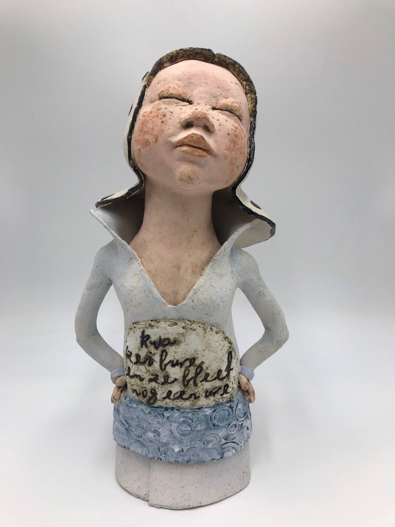 K&ouml;nnte beinhalten: Eine Keramikskulptur einer Frau mit geschlossenen Augen, die einen wei&szlig;-schwarz gemusterten Hut und ein hellblaues Kleid tr&auml;gt. Die Skulptur hat ein Schild auf der Vorderseite mit der Aufschrift "Kya er is burg en ze bleef nog een wee".