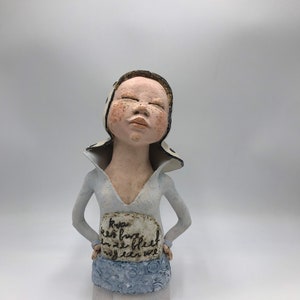K&ouml;nnte beinhalten: Eine Keramikfigur einer Person mit geschlossenen Augen, die einen wei&szlig;-schwarzen Hut und ein hellblaues Kleid tr&auml;gt. Die Figur h&auml;lt ein Schild mit der Aufschrift "Kiva keri hug in ze bleef nog eden ure".