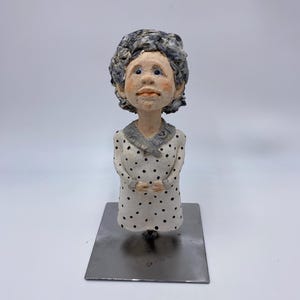 May include: A handcrafted ceramic sculpture of a woman with gray curly hair, wearing a white dress with black polka dots. The sculpture has a neutral skin tone and is mounted on a rectangular metal base.