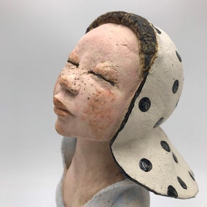 K&ouml;nnte beinhalten: Eine Keramikskulptur eines Frauenkopfes und -oberk&ouml;rpers. Die Frau tr&auml;gt einen wei&szlig;en Hut mit schwarzen Punkten. Ihre Augen sind geschlossen und sie hat einen friedlichen Ausdruck.