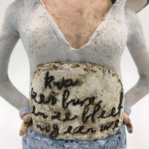 K&ouml;nnte beinhalten: Eine Keramikskulptur einer Frau in einem hellblauen Kleid mit wei&szlig;em Kragen. Das Kleid hat einen tiefen V-Ausschnitt und einen Rock mit einem kreisf&ouml;rmigen Muster. Der Torso der Frau ist mit einer wei&szlig;en Plakette mit schwarzer Schrift verziert, auf der steht "Kva en is bure en ze bleef nog een...