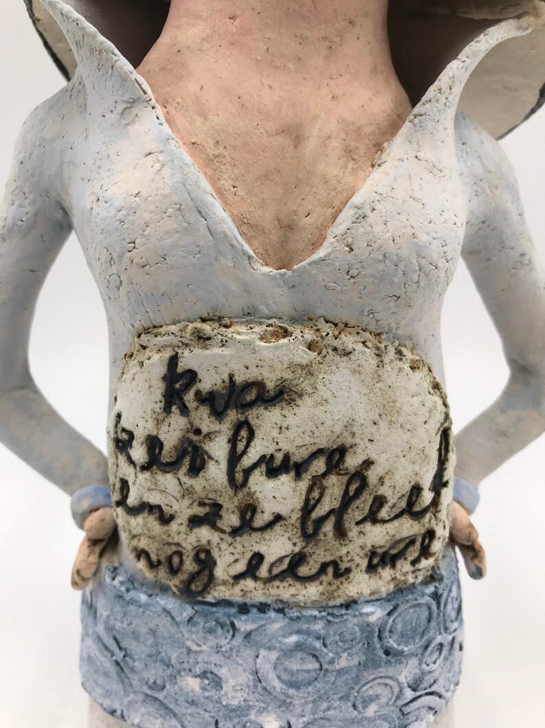 K&ouml;nnte beinhalten: Eine Keramikskulptur einer Frau in einem hellblauen Kleid mit wei&szlig;em Kragen. Das Kleid hat einen tiefen V-Ausschnitt und einen Rock mit einem kreisf&ouml;rmigen Muster. Der Torso der Frau ist mit einer wei&szlig;en Plakette mit schwarzer Schrift verziert, auf der steht "Kva en is bure en ze bleef nog een...