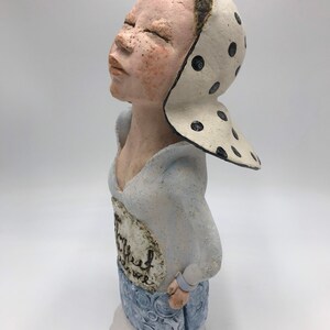 K&ouml;nnte beinhalten: Eine Keramikfigur einer Frau mit geschlossenen Augen, die ein wei&szlig;-schwarz gepunktetes Kopftuch und ein blau-wei&szlig; gemustertes Kleid tr&auml;gt. Das Kleid hat einen wei&szlig;en Kreis mit dem Wort "Thankful" darauf.