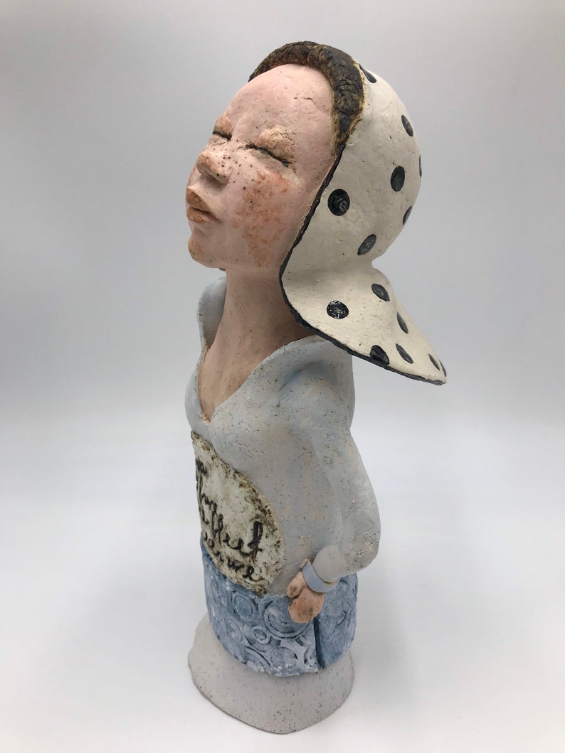 K&ouml;nnte beinhalten: Eine Keramikfigur einer Frau mit geschlossenen Augen, die ein wei&szlig;-schwarz gepunktetes Kopftuch und ein blau-wei&szlig; gemustertes Kleid tr&auml;gt. Das Kleid hat einen wei&szlig;en Kreis mit dem Wort "Thankful" darauf.