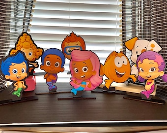 May include: A set of eight colorful cartoon character cutouts, including a yellow fish, a purple fish, a blue fish, a brown fish, a yellow character with a red hat, a pink character with a blue tail, a purple character with a yellow star, and a blue character with a green tail. The characters are standing on black bases.