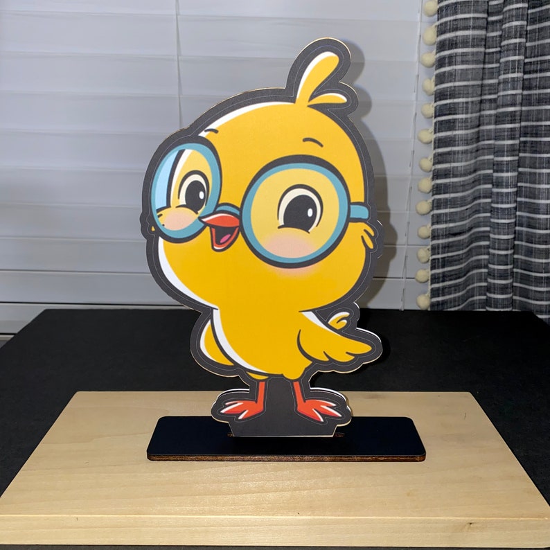 May include: A yellow chick wearing blue-rimmed glasses with a black stand. The chick has a red beak and orange feet.