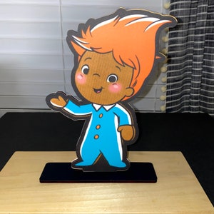 May include: A cartoon character cutout of a boy with orange hair wearing a blue jumpsuit with white accents. The cutout is standing on a black base.