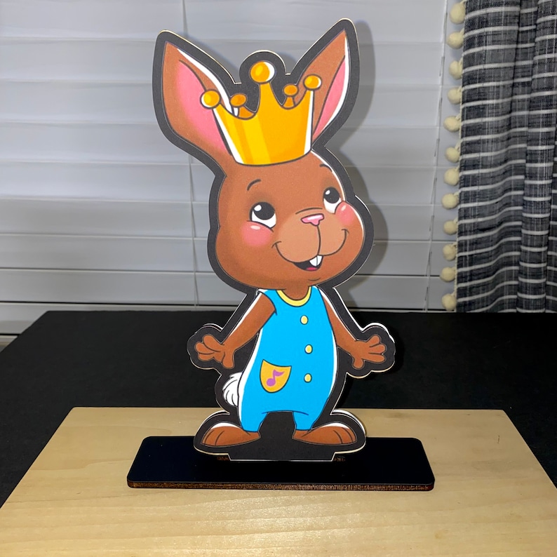 May include: A brown bunny wearing a yellow crown and a blue jumpsuit with a musical note on the pocket. The bunny is standing on a black base.