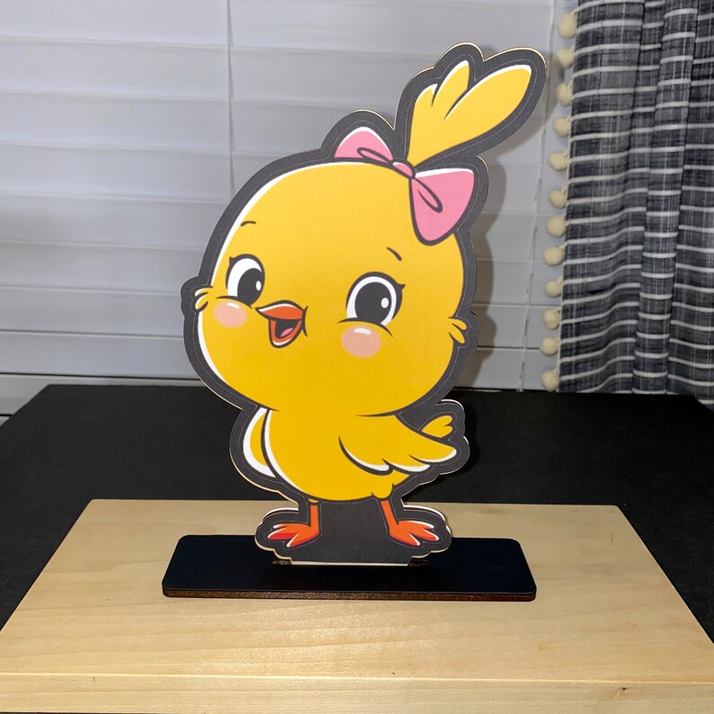 May include: A yellow chick with a pink bow, standing on a black base. The chick has a happy expression and is looking to the right. The chick is a cartoon character.