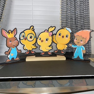 May include: Five colorful cardboard cutouts of cartoon characters. A brown bunny wearing a blue and white outfit and a crown, a yellow chick with glasses, a yellow chick with a pink bow, a yellow chick with a blue sailor hat, and a brown troll wearing a blue and white outfit. The cutouts are standing on a wooden surface.