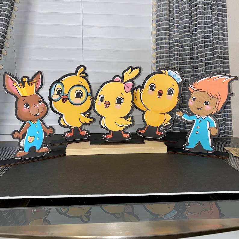 May include: Five colorful cardboard cutouts of cartoon characters. A brown bunny wearing a blue and white outfit and a crown, a yellow chick with glasses, a yellow chick with a pink bow, a yellow chick with a blue sailor hat, and a brown troll wearing a blue and white outfit. The cutouts are standing on a wooden surface.