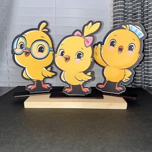 May include: Three yellow chick figurines with different expressions. The chick on the left is wearing glasses, the chick in the middle has a pink bow, and the chick on the right is wearing a blue and white sailor hat. The chicks are standing on wooden bases.