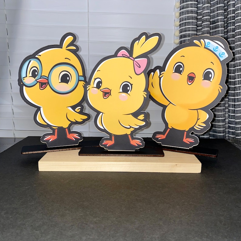 May include: Three yellow chick figurines with different expressions. The chick on the left is wearing glasses, the chick in the middle has a pink bow, and the chick on the right is wearing a blue and white sailor hat. The chicks are standing on wooden bases.