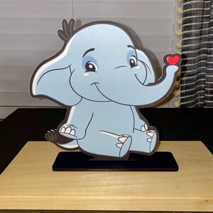 May include: A blue elephant figurine with a red heart on its trunk. The elephant is sitting on a wooden base.