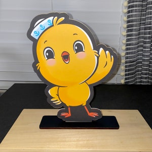 May include: A yellow chick wearing a blue and white sailor hat with an anchor design. The chick is waving with its right wing and has a happy expression on its face. The chick is standing on a black base.