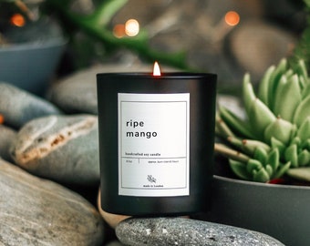 May include: A black soy candle with a white label that reads "ripe mango" and "handcrafted soy candle". The candle is sitting on a pile of gray and white stones.
