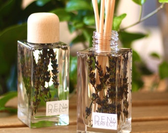 Puede incluir: Dos botellas de vidrio transparente con tapas de madera y flores de lavanda en su interior. Las botellas están llenas de un líquido transparente y tienen palitos de madera insertados en ellas. Las botellas están etiquetadas con "DENA PERFECT".