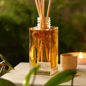 May include: A clear glass bottle diffuser with wooden sticks and a label that reads "DENA PERFECT". The bottle is filled with a golden liquid and dried flowers. The diffuser is sitting on a white surface with a wooden candle holder and a lit candle in the background.