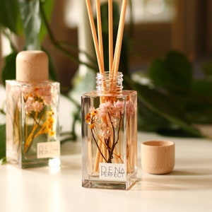 Puede incluir: Botellas difusoras cuadradas de vidrio transparente con tapas y varillas de madera. Las botellas contienen flores secas y el texto "DENA PERFECT". Una pequeña taza de madera está al lado de una botella. Hojas verdes en el fondo.