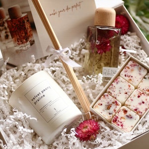 May include: A white gift box filled with white shredded paper. The box contains a white candle with a label that reads "velvet peony praline", a clear glass bottle with a wooden lid and dried flowers inside, and a white box with six white wax melts with pink flower petals inside. The box also includes a set of wooden diffuser sticks.